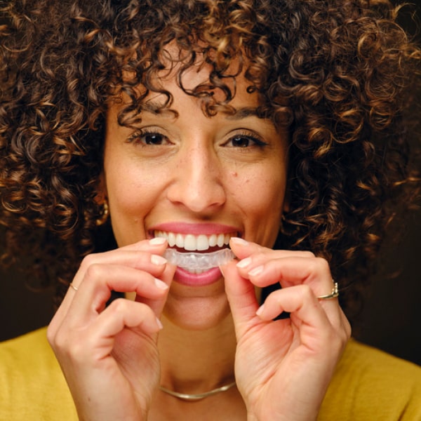 A female with curly hair smiles and holds a clear aligner